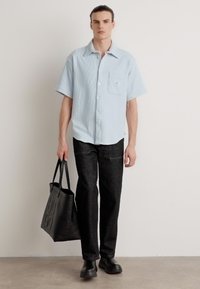Camicia azzurra a maniche corte con bottoni e taschino sul petto, abbinata a pantaloni scuri e texture e una borsa tote nera con logo impresso.