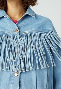 Světle modrá džínová bunda s třásněmi na přední straně, kovovými knoflíky a malou značkovou nášivkou na levé straně.