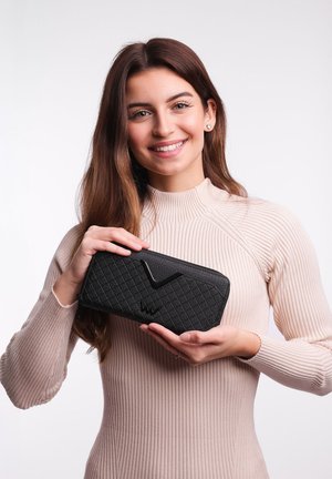 Black quilted wallet with a textured surface, featuring a prominent black logo and zip closure, held by a person wearing a ribbed beige top.