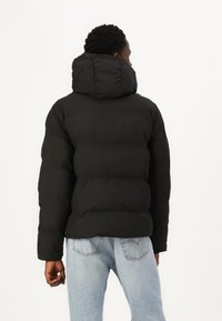 Sort pufferjakke med høj krave, prydet med et quiltet mønster og hætte. Bæres over lyseblå jeans. Glat overflade.
