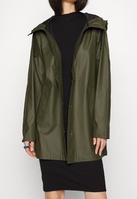 Giacca impermeabile verde oliva con cappuccio, chiusura con bottoni automatici e tasche laterali, abbinata a un vestito nero a coste. Texture liscia.