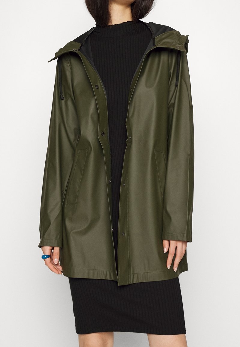 Giacca impermeabile verde oliva con cappuccio, chiusura con bottoni automatici e tasche laterali, abbinata a un vestito nero a coste. Texture liscia.
