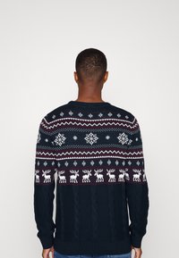 Stickad tröja med en mörkblå bas, snöflinge- och renmönster i vitt och vinröd, samt en kabelstickad textur på den nedre delen.