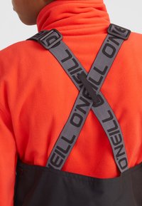 Čierne overal s šedými ramennými popruhmi O'Neill, prekríženými na chrbte, nosené cez jasne oranžový fleece top. Viditeľný textúrovaný detail látky.