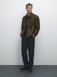 Braune Lederjacke mit Knopfverschluss, kombiniert mit grauen weiten Hosen und dunklen Schuhen, vor einem schlichten hellen Hintergrund.