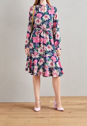 Kvinde iført en mørkeblå blomsterprint midi-kjole med lyserøde, hvide og lilla blomster, en rund udskæring og lyserøde højhælede sko.