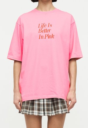 Osoba nosząca jasnoróżową, oversize T-shirt z tekstem "Life Is Better In Pink" i brązową plisowaną spódnicę w kratę.