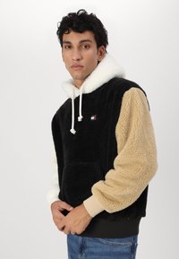 Hanorac din fleece, cu corp negru, glugă crem și mâneci bej. Dotat cu o buzunar frontal și sireturi ajustabile. Manșete tricotate în contrast.