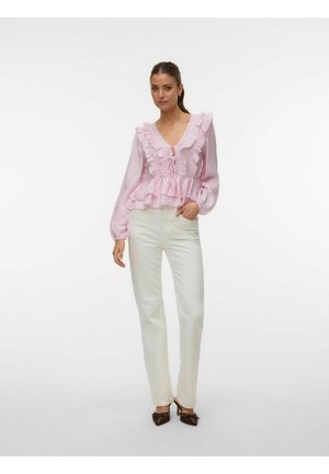 Femme portant un chemisier rose clair à volants avec des manches longues, un pantalon blanc taille haute et des talons pointus marron, debout devant un fond uni.