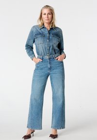 Spijkershirt en high-waisted, wijde jeans in lichtblauw. Het shirt heeft twee borstzakken en een knoopsluiting. Model draagt polkadot hakken.