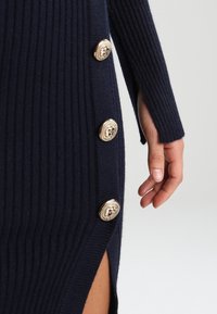 Marineblaues, geripptes Kleid mit goldenen Löwenkopfknoepfen an der Seite und einem schulterfreien Schlitz, aus strukturiertem Strickmaterial gefertigt.
