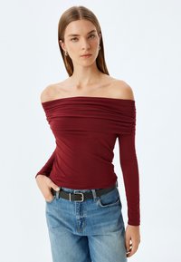 Bordeaux off-shoulder top med samlet stof; lange ærmer; parret med højtaljede blå jeans og et sort bælte. Glat tekstur.