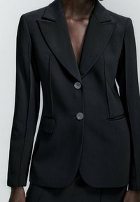 Blazer noir cintré avec un col à revers cranté, une fermeture à deux boutons et des poches latérales. Tissu lisse avec des coutures structurées qui mettent en valeur une silhouette ajustée.
