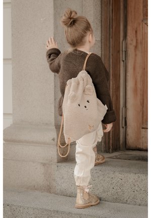 Bežan ruksak s vezicama, mekan i pahuljast, s elementima medvjeda, uključujući dvije uši i lice. Nosilo ga je dijete u smeđem džemperu i bijelim hlačama. Sjajne čizme.