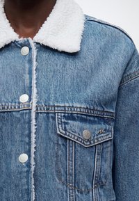 Gros plan d'une veste en jean avec doublure sherpa blanche au col et poche poitrine boutonnée sur une personne à la peau foncée.