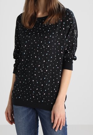 Woman wearing black long-sleeve top with small pink, white, and blue heart patterns and blue jeans against a plain background.