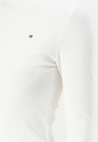 Weißes, geripptes Langarmoberteil mit kleinem gesticktem Logo. Der Stoff hat eine weiche Textur mit subtilen horizontalen Linien.