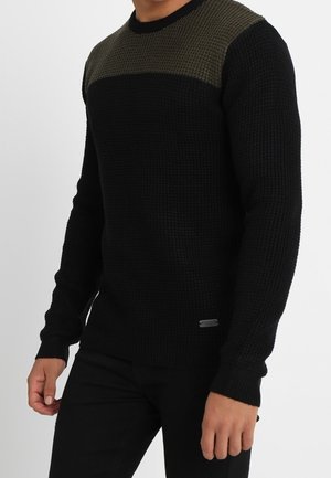 Pull en tricot noir avec un design bicolore, doté d'une section supérieure texturée en vert olive ; poignets et ourlet côtelés ; détail logo discret.