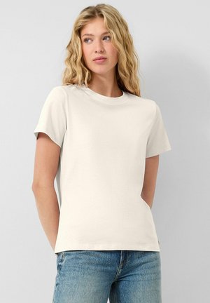 Blondynka z falującymi włosami, ubrana w jednolity, kremowo-biały, krótkorękawowy T-shirt i niebieskie dżinsy, stoi na tle jasnoszarego tła.