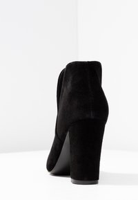 Bota de tobillo de ante negro con punta redondeada y tacón grueso. La parte trasera presenta un acabado suave con un sutil detalle de costura.
