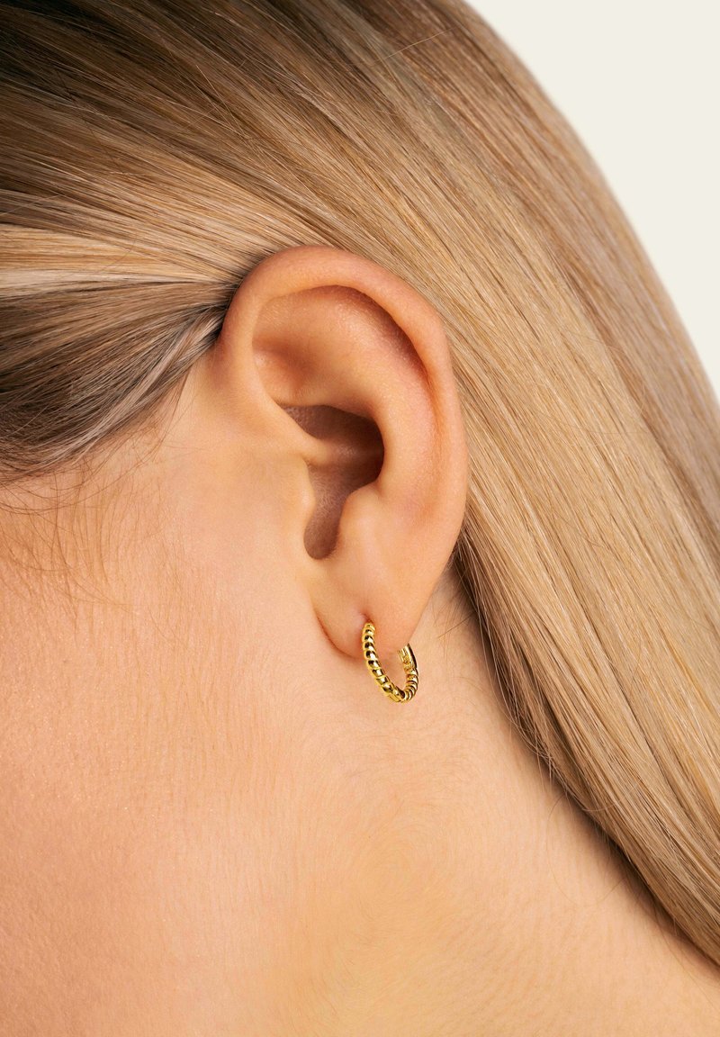 Pendiente de aro dorado con un diseño texturizado y retorcido. Visible en una oreja, destacando su forma circular y acabado brillante.