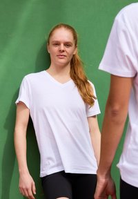 T-shirt bianca a collo a V con maniche corte, realizzata in tessuto liscio, indossata con pantaloni corti neri su uno sfondo verde uniforme.