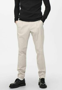Pantalons chinos beige avec une coupe décontractée, présentant des plis à l'avant et des poches latérales, associés à des chaussures noires à lacets.