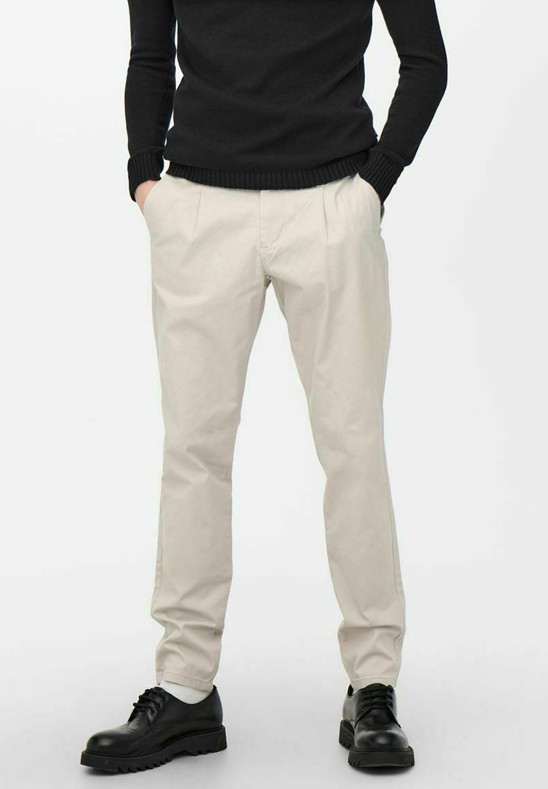 Pantalons chinos beige avec une coupe décontractée, présentant des plis à l'avant et des poches latérales, associés à des chaussures noires à lacets.