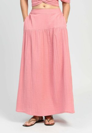 Femme portant une longue jupe rose fluide avec des poches et des sandales plates dark à fines lanières, debout devant un fond clair uni.