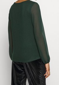 Grünes Langarmshirt mit festem Stoff am Rücken und transparenten Ärmeln. Die Ärmel sind am Bündchen leicht gerafft, was dem Design Struktur verleiht.