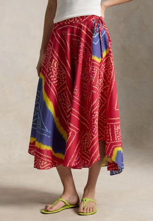 Žena oblečená v červenej zaviazanej sukni so žltými a modrými vzormi, bielom tričku a limetkovo zelených žabkách, stojaca s jednou rukou na boku.