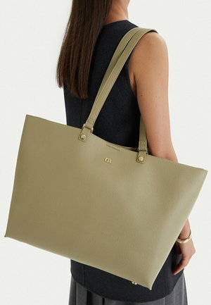 Mujer con vestido negro sin mangas lleva un gran bolso tote de textura en verde oliva claro sobre el hombro, con fondo liso.