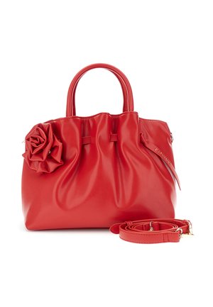 Červená kožená kabelka s nařasenými detaily, velkou růžovou ozdobou na boku, dvěma uchy a odnímatelným ramenním popruhem stočeným vedle ní.