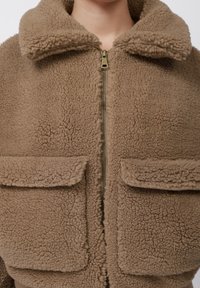 Barna sherpa polárdzseki túlméretezett gallérral, elöl fém cipzárral és két nagy fedeles zsebbel a mellkason.