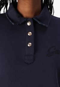 Marineblauwe polo met een klassieke kraag, drie gouden knopen en geborduurde accenten. Glad textuur met contrasterende details op de kraag.
