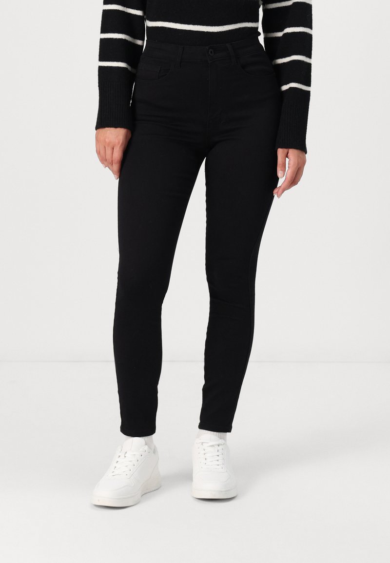 Zwarte skinny broek met een middellange taille, gemaakt van een soepele, rekbare stof, gecombineerd met witte sneakers voor contrast. Gestreepte trui er bovenop.