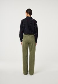 Vrouw met een knot in haar haar, gekleed in een zwart shirt met kleurrijke hartpatronen en high-waisted olijfgroene jeans, staande met haar rug naar de camera op een witte achtergrond.