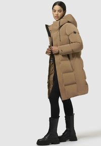 Lang, puffy brun frakke med hætte, med sorte accenter og en quiltet mønster. Båret over sorte leggings og snørebåndssorte støvler.