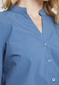 Lichtblauw, knoopshirt met een Mandarin-kraag, een gladde textuur en subtiele stitchingdetails. De knopen zijn rond en transparant.