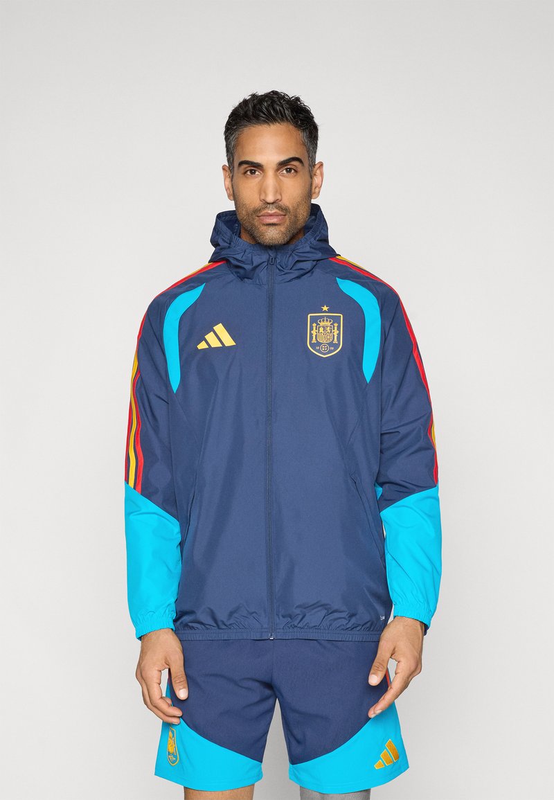 Námornícka športová bunda s tyrkysovými rukávmi, červenými a žltými pruhmi a zlatým erbom Španielska; doplnená o zodpovedajúce námornícke a tyrkysové šortky.