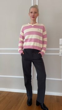 Person standing indoors wearing a pink and white striped sweater, dark pants, and black heeled boots, with hands in pockets.