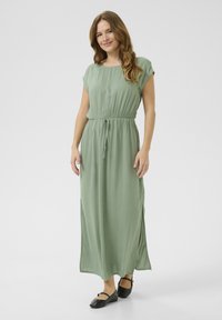 Maxi dress in soft, light green fabric with short sleeves, an elastic waist, and side slits; paired with black flat shoes.