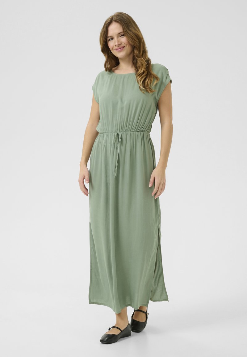 Maxi dress in soft, light green fabric with short sleeves, an elastic waist, and side slits; paired with black flat shoes.