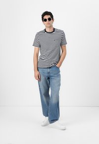 Striped short-sleeve t-shirt in black and white, paired with light blue straight-leg jeans and white sneakers. Model wears dark sunglasses.