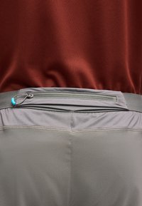 Gros plan sur un pantalon de sport gris avec une poche zippée horizontale à la taille et un haut bordeaux rentré à l’intérieur.