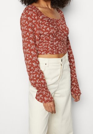 Mujer con un top corto de flores de manga larga color óxido combinado con pantalones de tiro alto en color blanco roto, sobre un fondo liso.