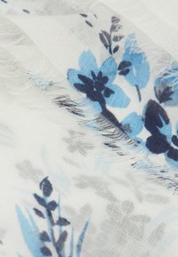 Écharpe florale avec des fleurs bleues et navy sur un fond blanc, réalisée en tissu léger avec des bords effilochés et une texture douce.