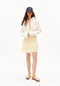 Chemise blanche à boutons, jupe jaune clair avec perforations circulaires et sandales oranges translucides. Une casquette marine complète la tenue.