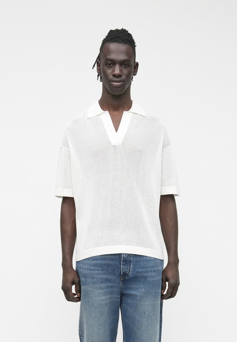 Biała dzianinowa koszulka polo z dekoltem w serek i krótkimi rękawami, z teksturowanym wzorem. W zestawie z jasnoniebieskimi dżinsami.