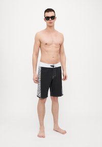 Sorte badeshorts har en hvit midjebånd med snøring og et dekorativt mønster langs sidene. Stoffet ser lett og hurtigtørkende ut.